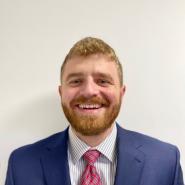 A man with a beard smiles at the camera. It is a headshot, and he is wearing a blue suit, white shirt, and red tie.