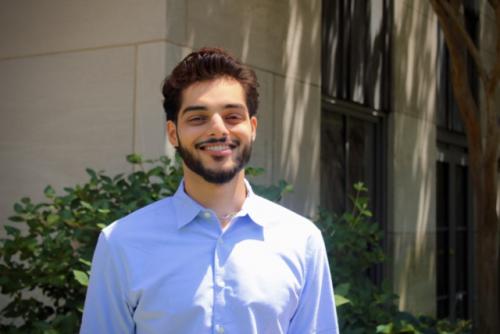 A man wearing a blue button up shirt is smiling at the camera. He has a beard. There is a plant behind him.