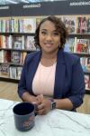 A woman with black hair smiles at the camera. She is wearing a blue blazer and a light pink shirt. There are bookshelves behind her.