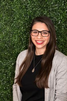 A woman with long brown hair is smiling at the camera. She is wearing a beige sweater over a black shirt and standing in front of green foliage. She has on red lipstick and dark glasses.