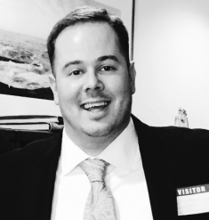 A black and white photo of a man smiling at the camera. He is wearing a suit and tie and has dark hair.
